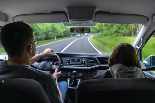 Driver and passenger inside a car on a tree-lined road, viewed from the back seat, illustrating a typical driving environment where road noise can occur.